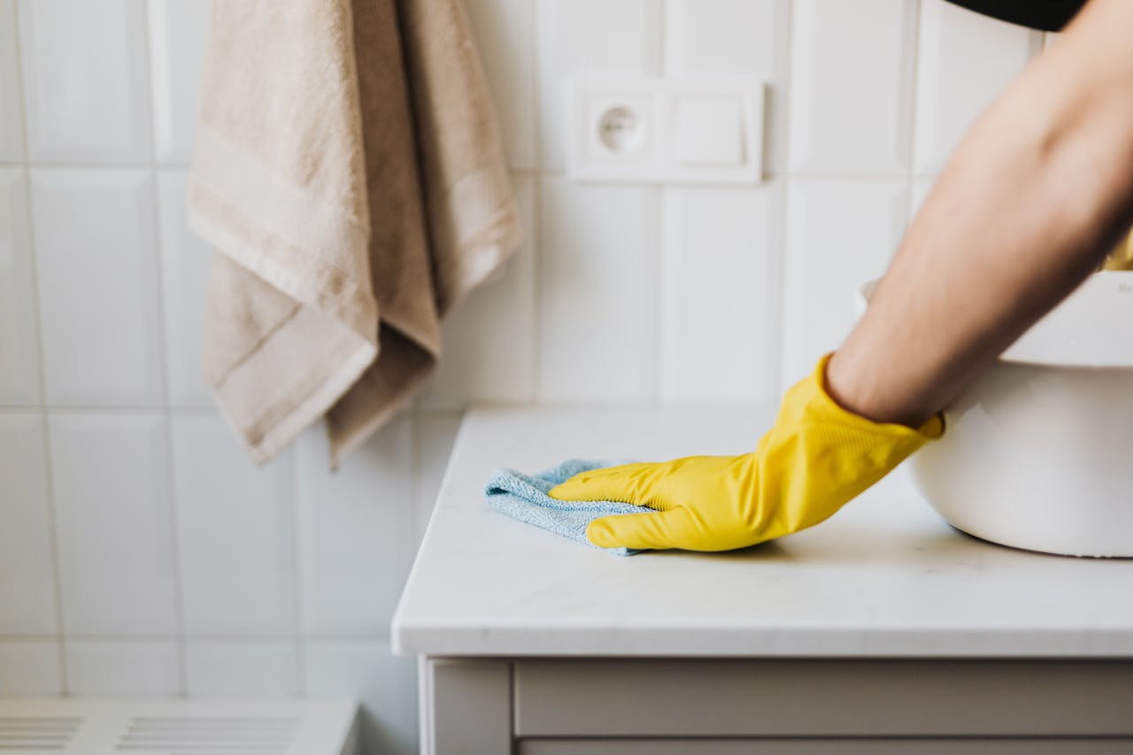 about-img Close-up of a person wearing yellow gloves wiping a bathroom counter with a cloth.